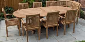 Teak outdoor dining table with eight matching teak chairs and a teal bench on a stone patio.