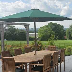 Teak patio dining set with a green parasol and a scenic green landscape in the background.